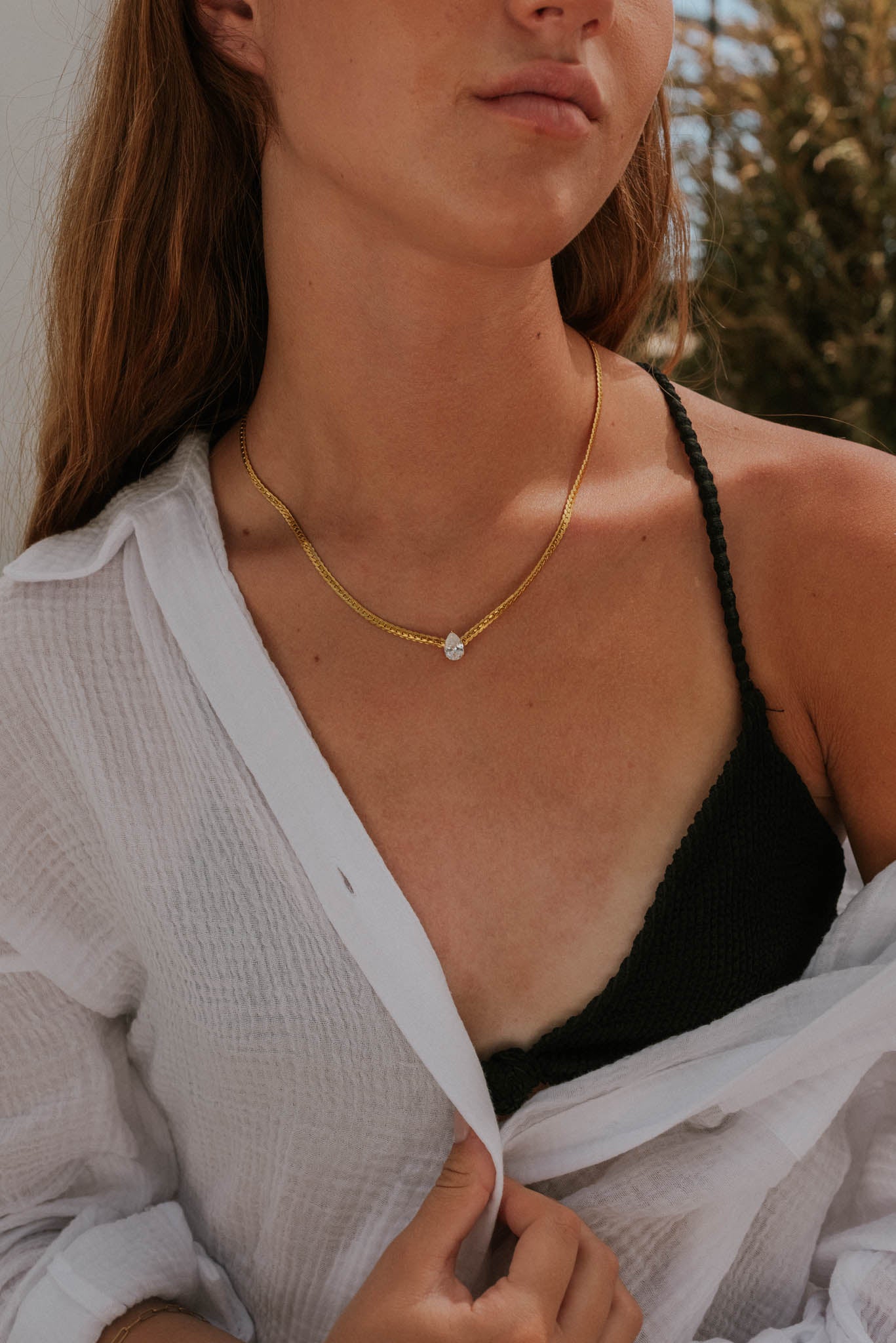 Woman wearing a gold necklace with a clear pendant, white shirt, and black top outdoors.