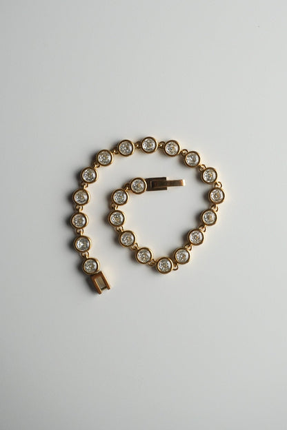 A gold tennis bracelet with round-cut clear stones, coiled on a plain light gray background.