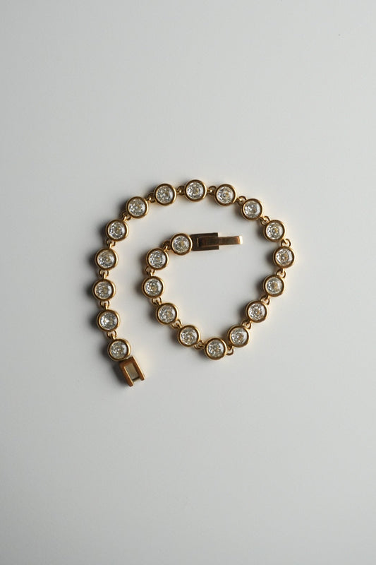 A gold tennis bracelet with round-cut clear stones, coiled on a plain light gray background.
