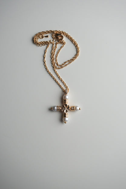 A gold cross pendant necklace with white pearl-like beads and a central clear rhinestone, resting on a light gray background.