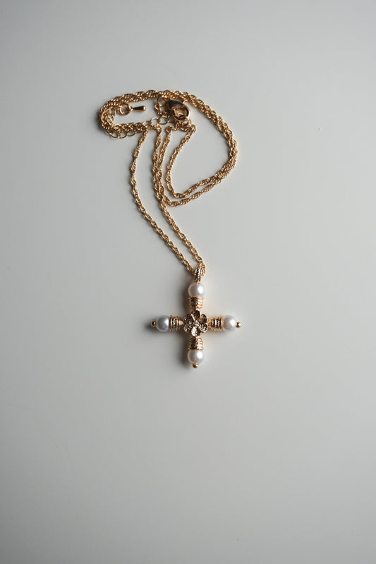 A gold cross pendant necklace with white pearl-like beads and a central clear rhinestone, resting on a light gray background.