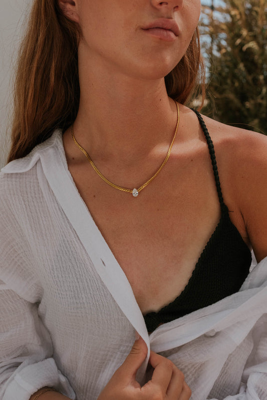 Woman wearing a gold necklace with a clear pendant, white shirt, and black top outdoors.