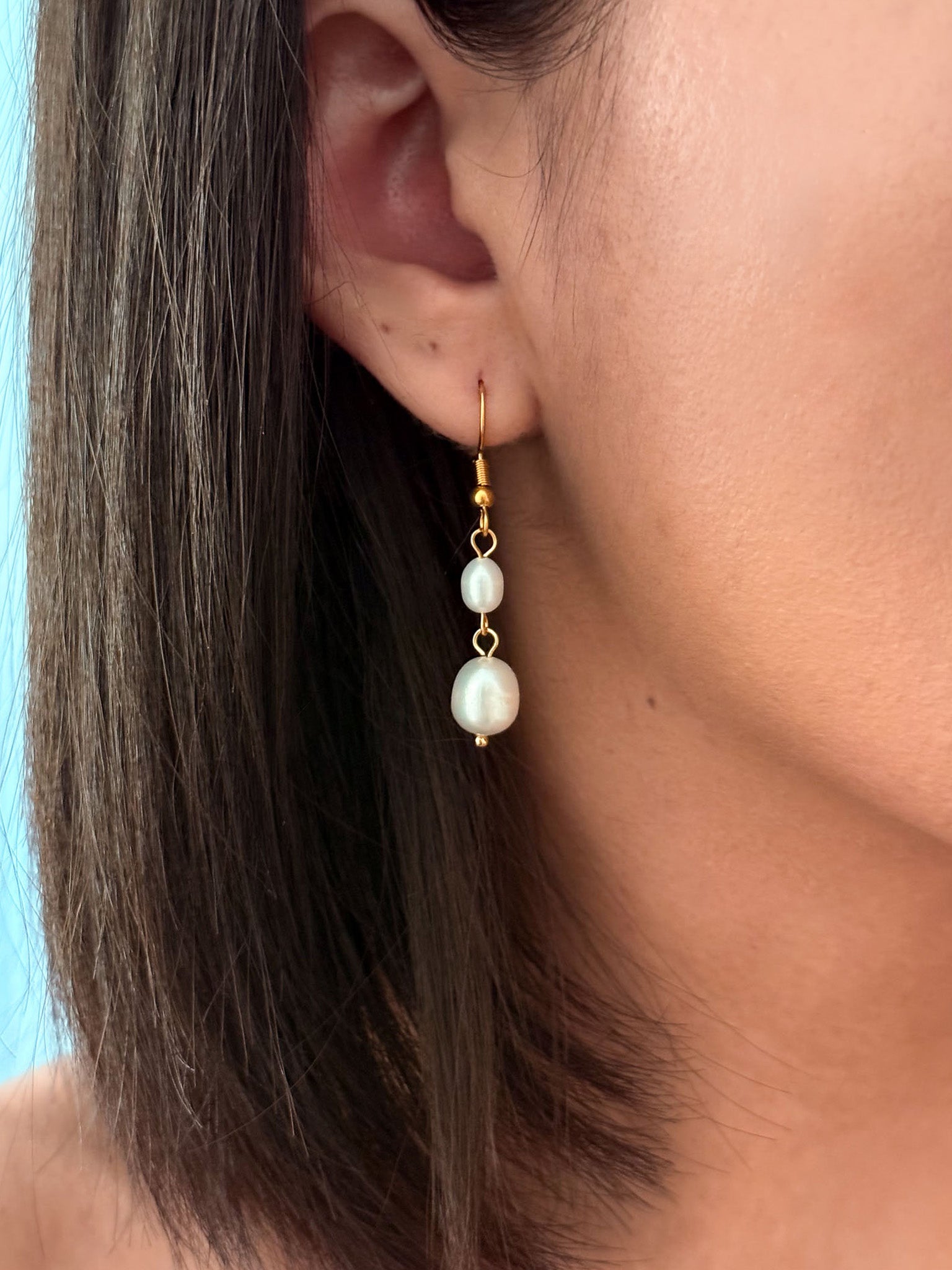 A close-up of a model's ear wearing a gold drop earring with two white pearl-like beads.